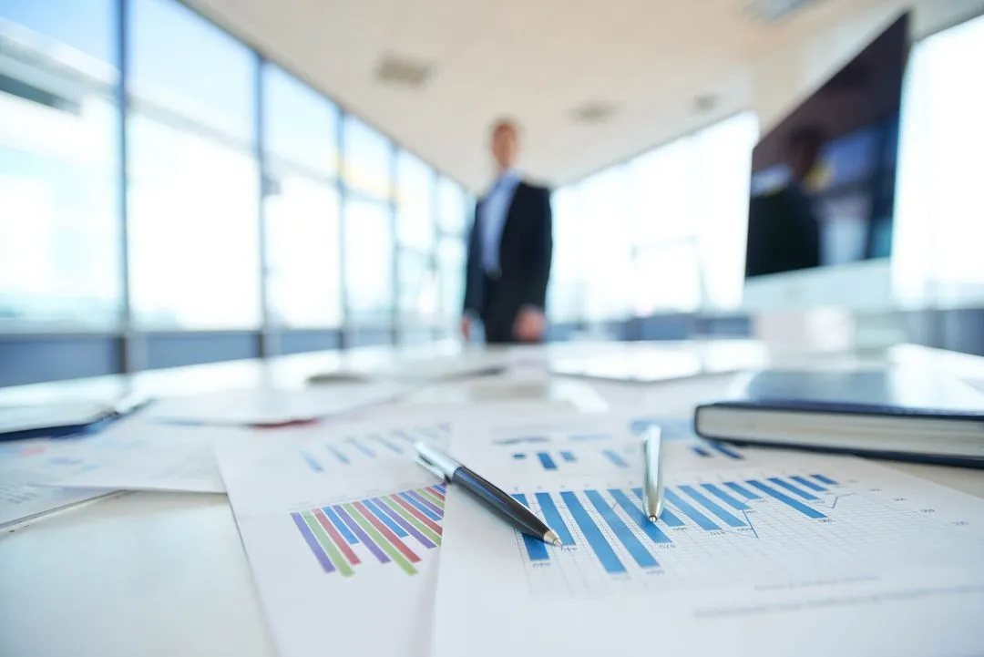 Business man in a top floor office with lots of windows standing at the end of a large table covered in paper work and computers with graphs 